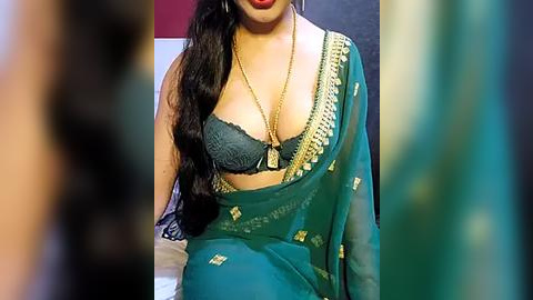 Media: Video of a South Asian woman in a green sari with gold embroidery, wearing a black lace bra, seated indoors.