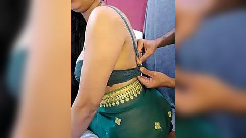 Media: Video of a South Asian woman with medium brown skin and long black hair, wearing a green sari with gold embroidery. A man in a blue shirt is adjusting her sari. Background includes a red wall and blurred figures.