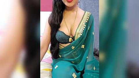 Media: Video of a South Asian woman in a green sari with gold embroidery, showcasing a teal lace bra. She has long black hair, fair skin, and wears a gold necklace. Background includes a pink wall and blurred figures.