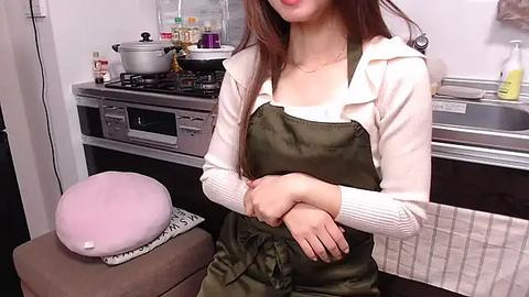 Media: Video of a woman in a white cardigan and olive green apron, standing in a modern kitchen with a pink hat on a stool, cooking pots on a stove, and white tiles.