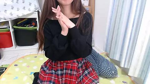 Media: Video of a young Asian woman with long brown hair, wearing a black top and red plaid skirt, kneeling on a polka-dotted green rug in a brightly lit room with white walls and a green curtain.