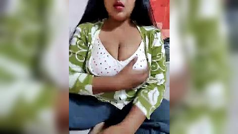 Media: A video shows a woman with medium brown skin, wearing a white polka-dotted top and a green floral robe, sitting with crossed arms, partially covering her breasts. The background is blurred.