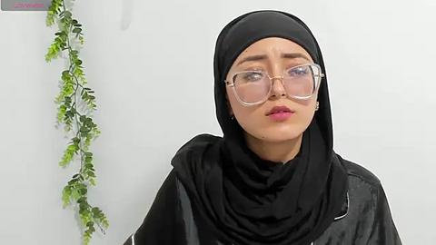 Media: Video of a light-skinned woman with a medium build, wearing a black hijab and clear glasses, in a plain white room adorned with green ivy.