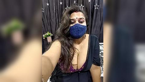 Media: Video of a South Asian woman with long dark hair, wearing a blue polka-dot face mask, purple lace bra, and black dress, seated in front of a starry backdrop.