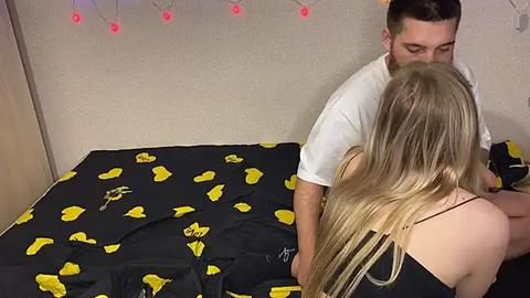 Media: Video of a man and woman on a bed with a black sheet adorned with yellow cartoon characters. The woman, with long blonde hair, wears a black tank top. The man, with short dark hair, kisses her neck.