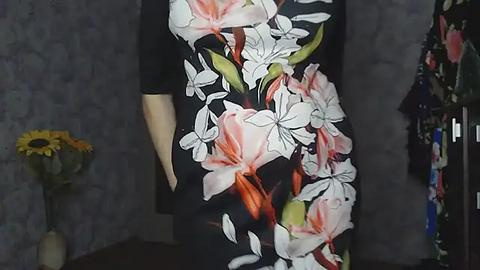 Media: Video of a person wearing a black dress adorned with a large, vibrant floral print featuring white and pink lilies with green leaves. Background includes a grey wall, a vase with sunflowers, and a partially visible rack of colorful clothes.