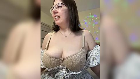 Media: Video of a smiling, middle-aged woman with glasses, fair skin, and brown hair, wearing a beige lace bra and off-shoulder floral dress, in a room with colorful string lights.