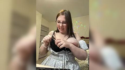 Media: A video shows a plus-size woman with glasses, light skin, and brown hair, wearing a black bra and a white floral dress, adjusting her bra in a cozy, softly lit bedroom.