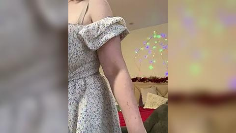 Media: Video of a fair-skinned woman with light brown hair in a floral dress, partially off-shoulder, standing in a cozy bedroom with Christmas lights and a bed adorned with a red blanket and pillows.