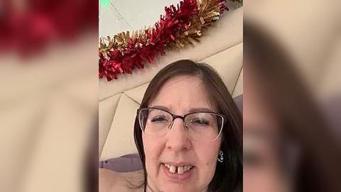 Media: Video of a middle-aged woman with glasses, smiling, wearing a beige sweater, against a background of a beige couch and festive red and gold tinsel garland.