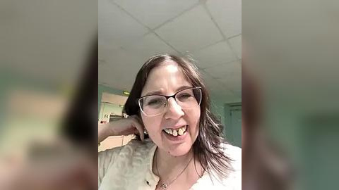 Media: A video of a Caucasian woman with shoulder-length brown hair, wearing glasses and a white shirt, smiling with braces. Background shows a blurred, tiled ceiling and green walls in a sterile, possibly hospital setting.