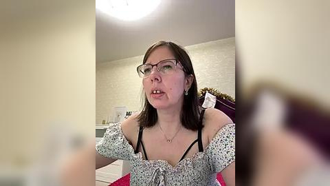 Media: Video of a white woman with glasses, dark hair, wearing a floral off-shoulder top, sitting on a bed, blurry background.