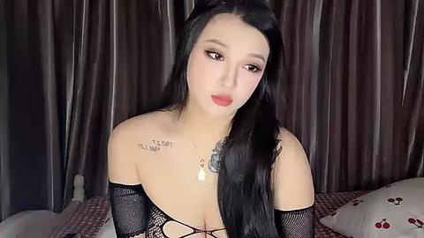 Media: Video of a fair-skinned woman with long black hair, wearing a black fishnet top, sitting on a bed with red and white checkered pillows, against dark brown curtains.