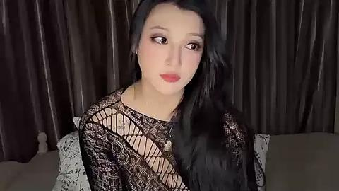 Media: Video of a pale-skinned, long-haired woman with black eye makeup and red lipstick, wearing a black, fishnet-patterned top. She gazes thoughtfully to the side, against dark, pleated curtains.