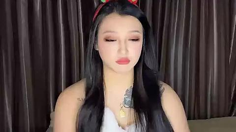 Media: Video of an East Asian woman with long black hair, wearing a red headband, red lipstick, and a white sleeveless top. She stands against dark, shiny curtains.