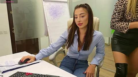 Media: Video of a young woman with long brown hair, light skin, and a slender build, wearing a light blue button-up shirt and blue jeans, sitting at a desk in an office.