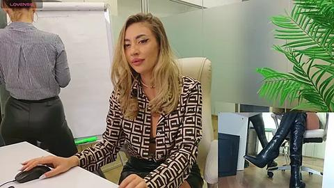 Media: Video of a blonde woman in a patterned blouse, seated at a white desk in an office. Background includes a potted plant, another person, and a greenboard.