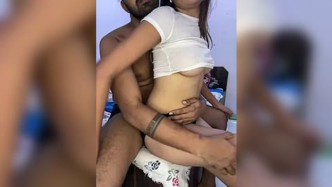 Media: Video of a nude man lifting a partially clothed woman in a white crop top and panties. Background shows a blurred bedroom with a bed and scattered objects.