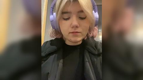 Media: Video of a young woman with short blonde hair, wearing oversized purple headphones, a black coat, and a black turtleneck, sitting on a public bus.