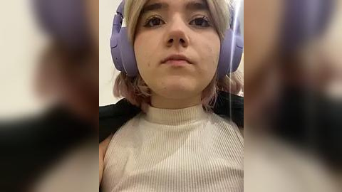 Media: Video of a young woman with fair skin, light blonde hair, and light purple headphones, wearing a ribbed beige turtleneck sweater, in a blurry background.