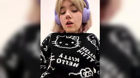 Media: Video of a young woman with light blonde hair wearing purple headphones and a black sweater with Hello Kitty and \"Kawaii\" text, looking relaxed against a blurred background.