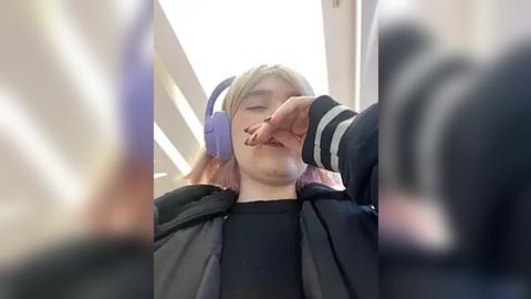 Media: Video of a young woman with pink hair, wearing headphones, lying on a train seat with a man's hand covering her mouth, blurred background.