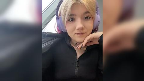 Media: A video of a young woman with light skin and blonde hair wearing large purple headphones, resting her chin on her hand. She has a neutral expression and is dressed in a black hoodie.