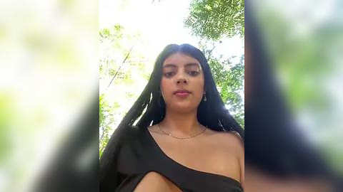 Media: Video of a young woman with long, straight black hair and a tan complexion, wearing a black off-shoulder top, standing in a sunlit forest with green foliage in the background.