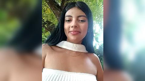 Media: Video of a young Latina woman with long black hair, wearing a white, off-shoulder knit dress, standing outdoors with blurred foliage in the background.