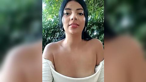 Media: A video of a young woman with medium brown skin and long black hair, wearing an off-shoulder white top, standing against a green leafy background.