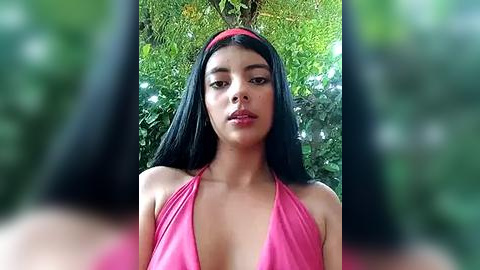 Media: Video of a young woman with long black hair, wearing a red bikini top, standing in a lush, green garden. Her expression is neutral, with a slight frown.