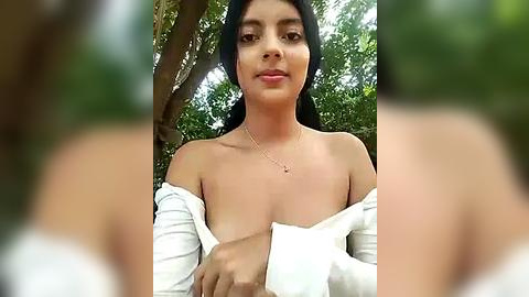 Media: A video of a young woman with medium brown skin, long black hair, and a slender physique, partially covered by a white off-shoulder top, standing outdoors in a lush green forest.