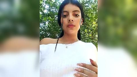 Media: Video of a young woman with medium brown skin, long black hair, and a white off-the-shoulder top, standing against a green leafy background.