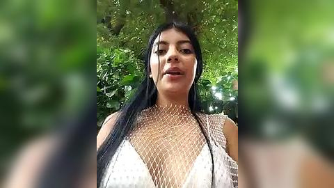 Media: Video of a dark-haired woman with medium skin tone, wearing a revealing, white mesh top, standing outdoors amidst lush green foliage. Her expression is neutral, and she appears to be in a relaxed, natural setting.