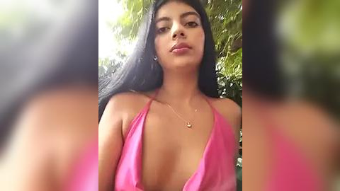 Media: Video of a young woman with long black hair, medium skin tone, wearing a bright pink halter top that reveals cleavage, standing outdoors against a green leafy background.