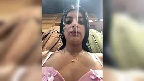 Media: A video of a young woman with medium brown skin and straight black hair, wearing a pink bra with floral details and a gold necklace. She is sitting on a wooden bed with a blurred background.