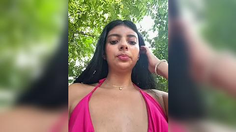 Media: Video of a young woman with medium brown skin and long black hair, wearing a bright pink halter top, standing outdoors amidst green foliage. Her expression is neutral.