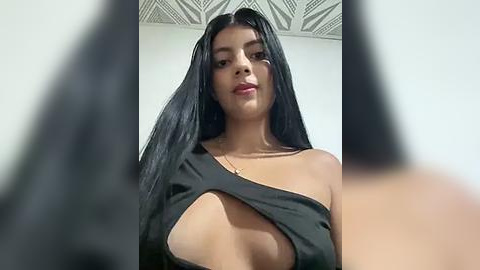 Media: Video of a young woman with long black hair, fair skin, and full lips, wearing a revealing black one-shoulder dress with a cut-out revealing her breast, set against a plain, white ceiling background.