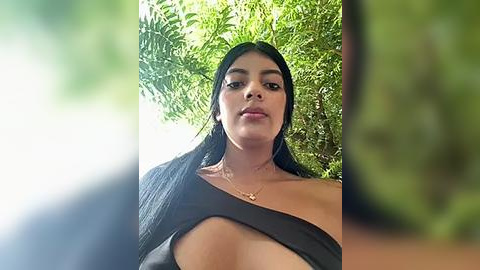 Media: Video of a young woman with long black hair, medium skin tone, and a slender physique, wearing a black off-shoulder top, standing against lush green foliage.