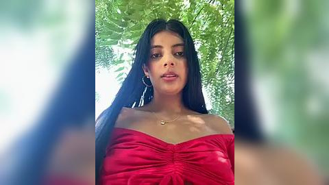 Media: A video of a young woman with long black hair and medium brown skin, wearing a red off-shoulder dress, standing in a lush, green forest. She has hoop earrings and a gold necklace.