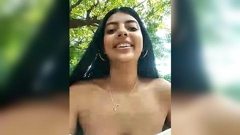 Media: Video of a topless young woman with long black hair, smiling, wearing a gold necklace, against a blurred green foliage background.