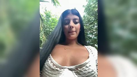 Media: Video of a young woman with long, straight black hair, medium brown skin, and a curvaceous figure, wearing a white, fishnet top revealing cleavage. Background features lush green foliage and palm trees, suggesting an outdoor tropical setting.