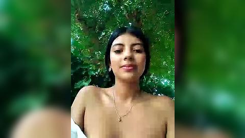 Media: Video of a topless woman with medium-sized breasts, tan skin, dark hair, and a necklace, standing outdoors amidst lush green foliage.