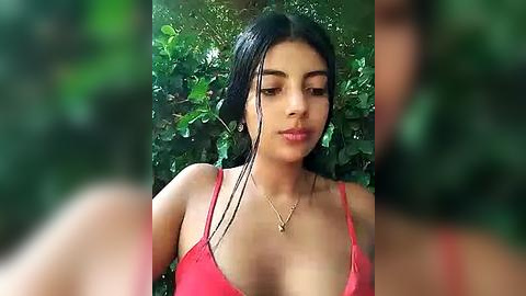 Media: Video of a young woman with olive skin and long black hair, wearing a red spaghetti-strap top, standing against a lush green background.