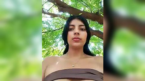 Media: Video of a young woman with long, dark hair and medium skin tone, wearing a strapless brown top, standing in a lush, green forest with sunlight filtering through the trees.