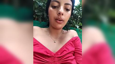 Media: A video of a young woman with medium skin tone, wearing a pink off-shoulder top and a delicate necklace, sitting outdoors against a green background with blurred foreground.