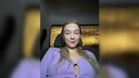 Media: Video of a young Caucasian woman with long blonde hair, wearing a lavender cardigan, seated in a dimly-lit interior with a textured golden backdrop.