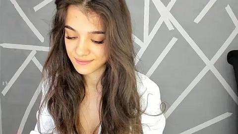 Media: Video of a young girl with long, wavy brown hair, wearing a white top, against a gray wall with geometric patterns. She appears serene and slightly smiling.