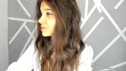 Media: Video of a young woman with long, wavy brown hair, wearing a white shirt, set against a geometric white patterned background.