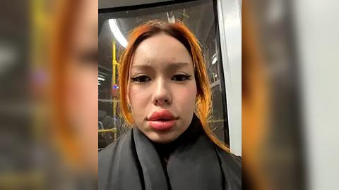 Media: Video of an Asian woman with fair skin and long, vibrant orange hair, wearing a black cape, standing inside a subway car with blurred, yellow seats and windows in the background.